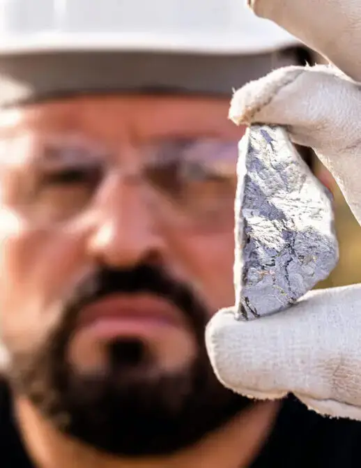 A miner in a hard hat holds a lump of silver ore up to the camera