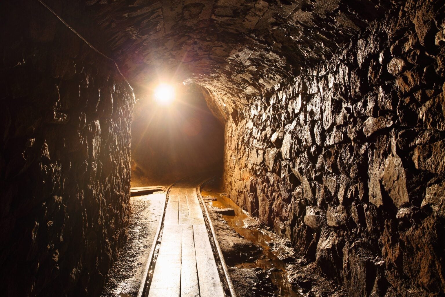 A lamp-lit mine tunnel with wooden walkway boards running toward a bright light