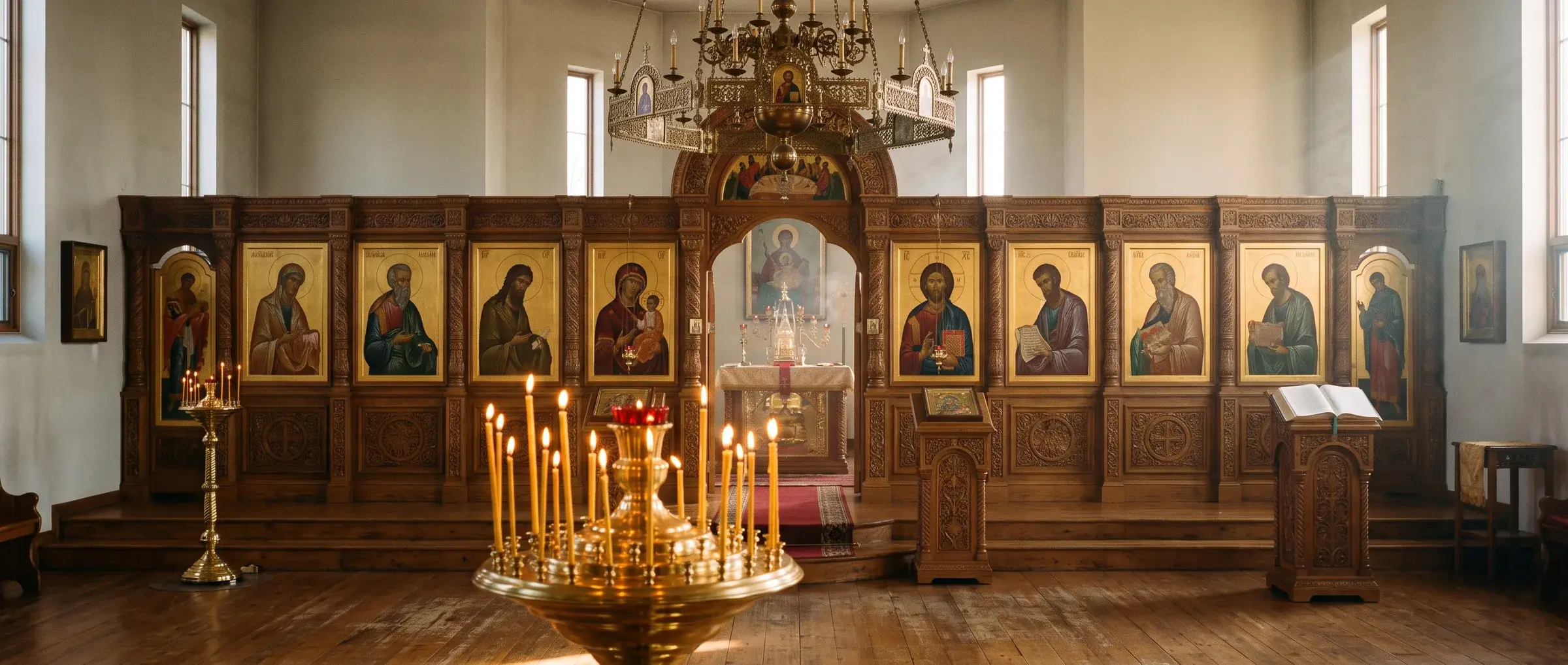 The interior of an Orthodox parish church in the evening: a carved wooden iconostasis with gilded icons, the Royal Doors open to the altar, and beeswax candles burning in a brass stand